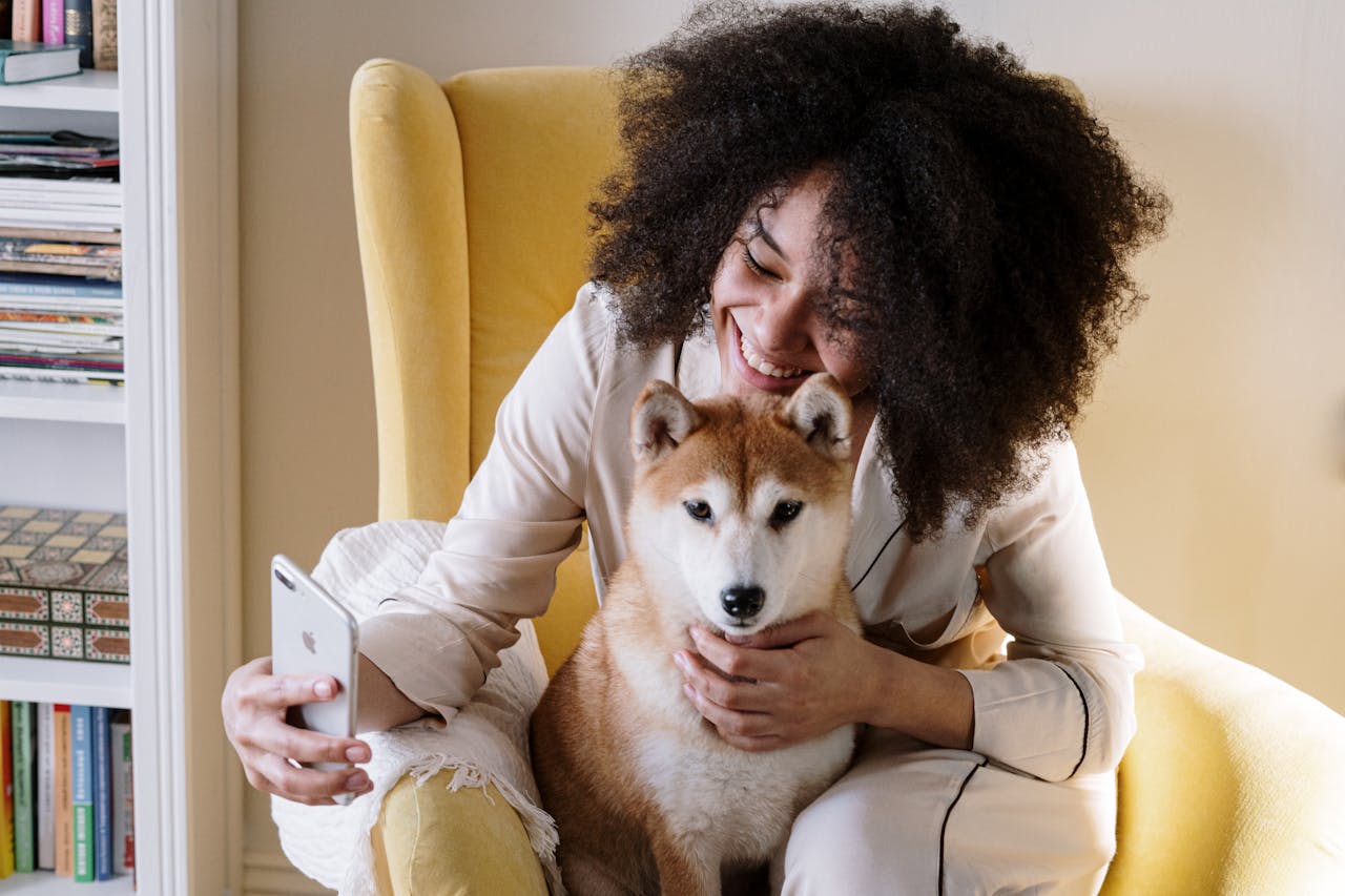 A joyful woman sitting on a yellow armchair taking a selfie with her Shiba Inu dog at home.