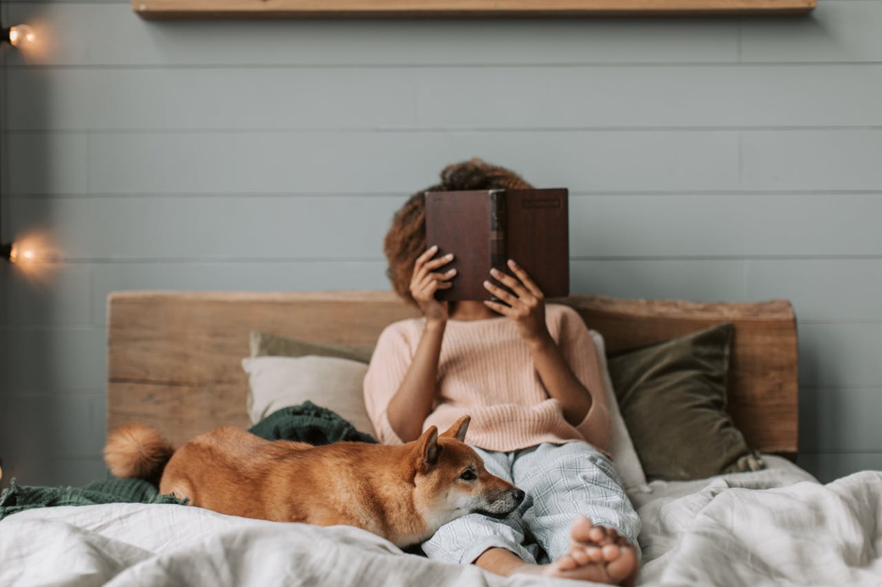 Woman relaxing on bed with book and Shiba Inu dog, cozy indoor scene.